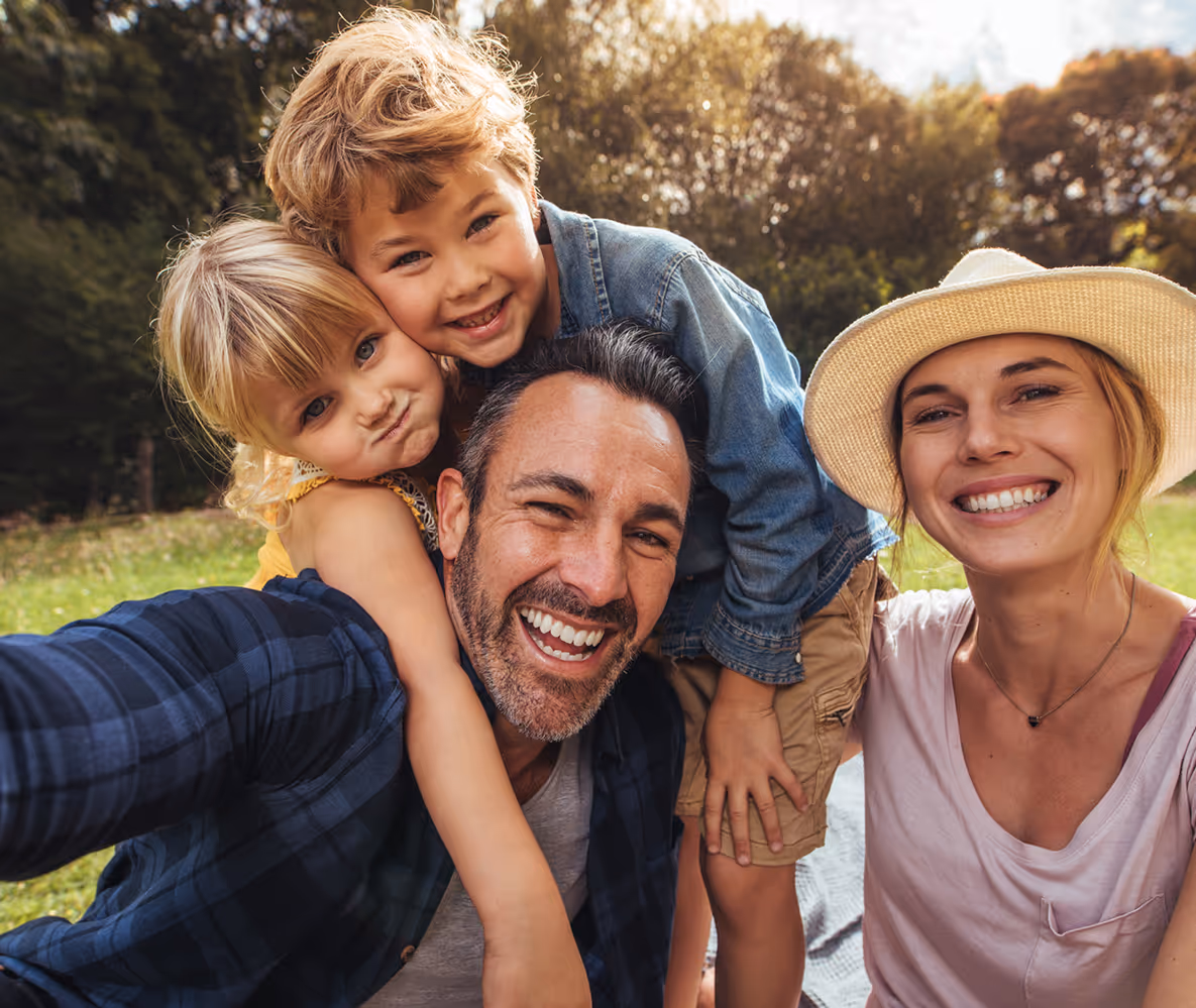 Smiling family of four taking a selfie outdoors on a sunny day with trees in the background.