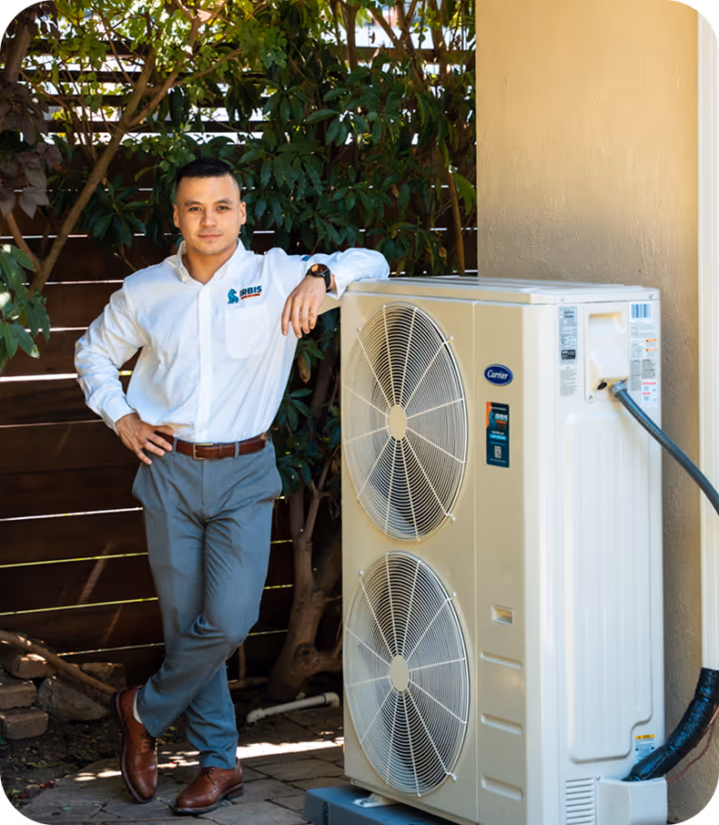 Man in white shirt and grey pants leaning on a large outdoor air conditioning unit with twin fans in a garden setting.