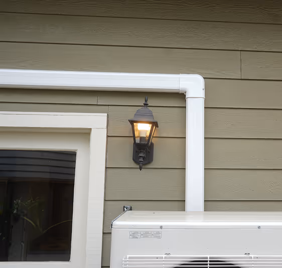 Outdoor wall light fixture turned on beside window with white piping and air conditioning unit.