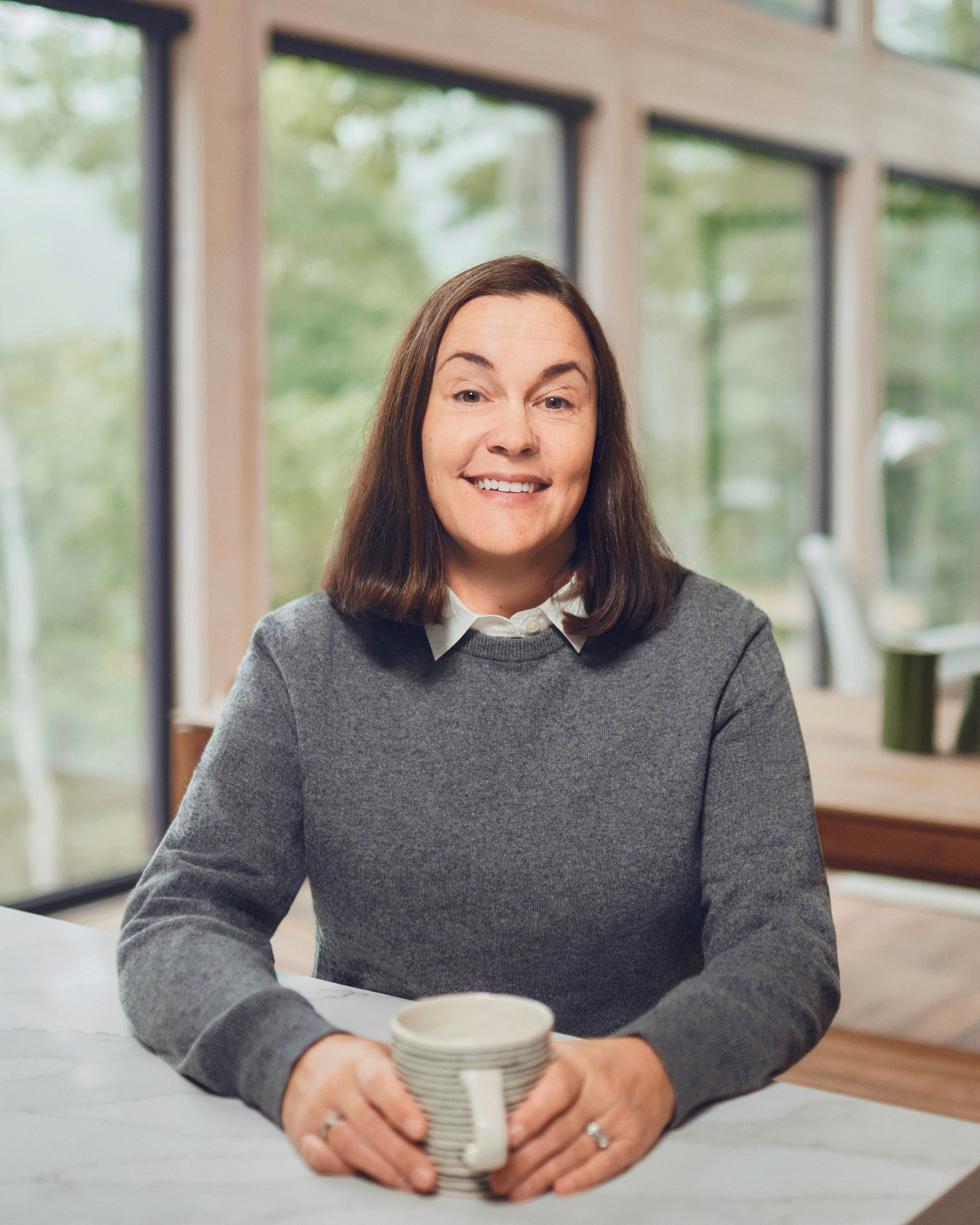 Brie Pio, founder and lead financial planner at Birch Point Wealth Management, smiling at a counter with a mug, tall windows behind her