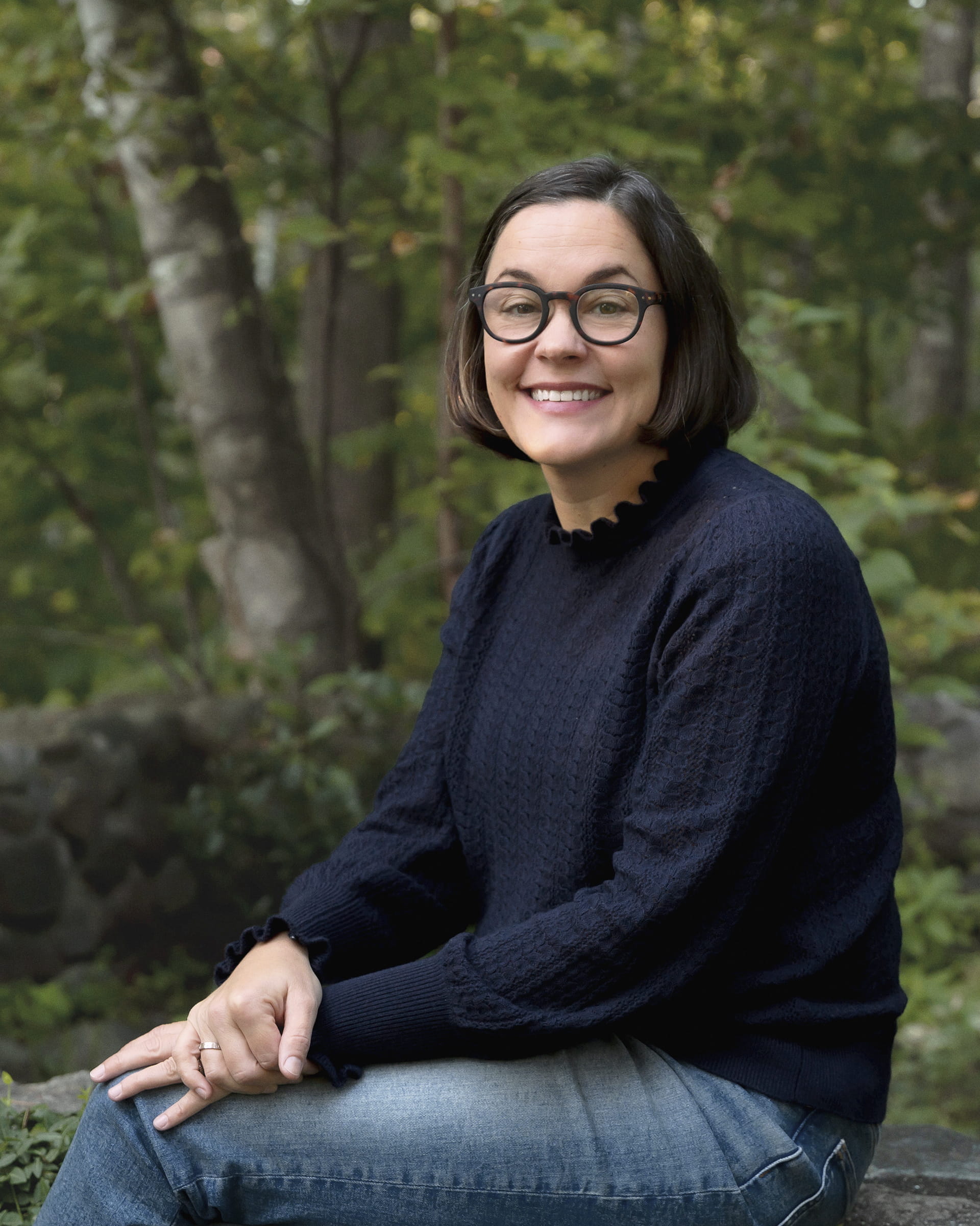 Brie Pio, founder and lead financial planner at Birch Point Wealth Management, seated on a stone wall in the woods