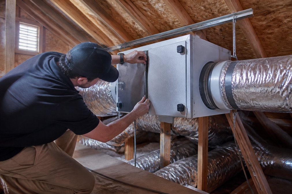 Professional technician in work attire installing attic-mounted air purification system with visible ductwork connections and HVAC integration in residential attic space with natural light from soffit vents