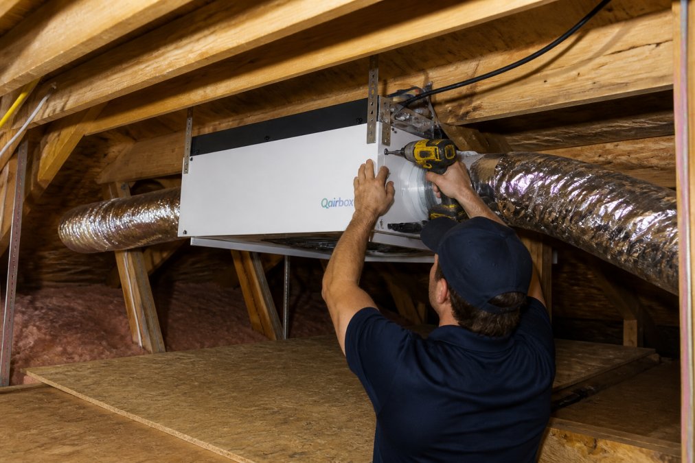 Professional technician installing an attic-mounted air purification system in a residential attic space, securing the unit to wooden rafters with visible ductwork connections and electrical wiring