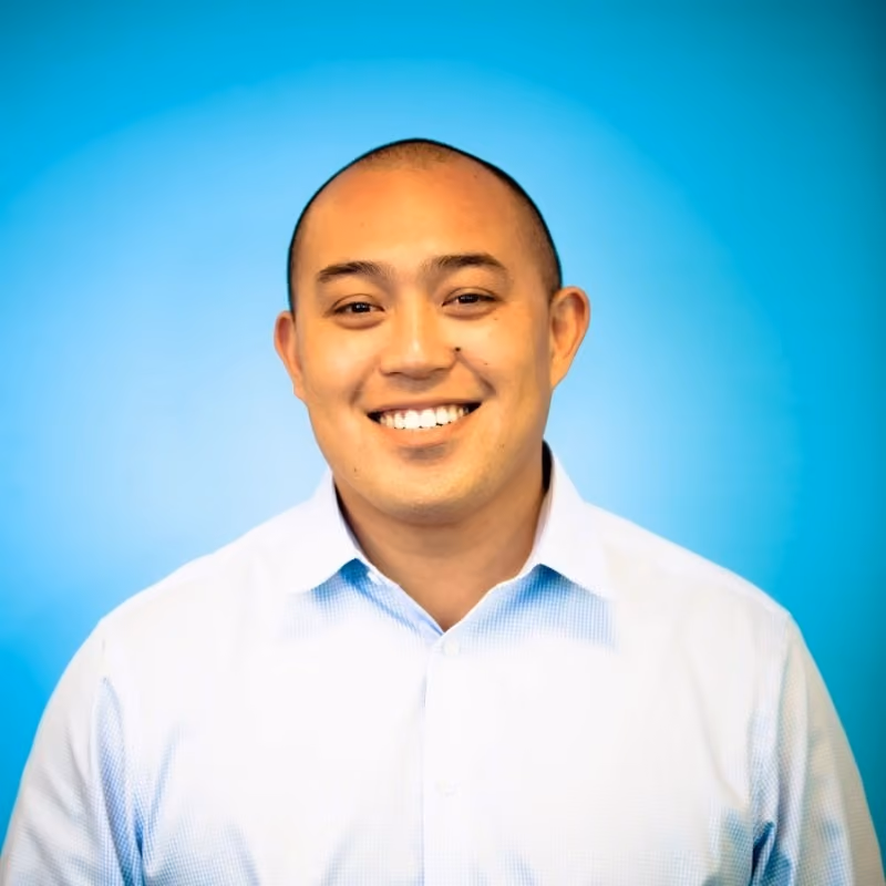 Smiling bald man wearing a light blue collared shirt against a blue gradient background.