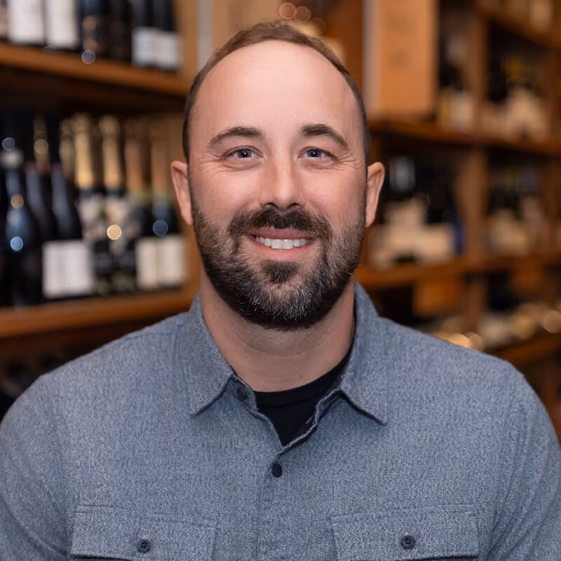 Smiling man with short beard and gray button-up shirt standing indoors with blurred bottles behind him.
