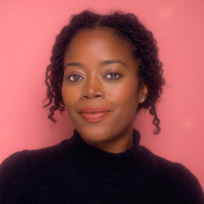 Close-up portrait of a woman with natural curly hair wearing a black turtleneck against a pink background.