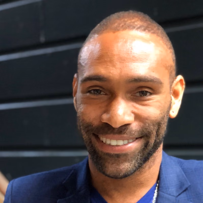 Smiling man with short hair and beard wearing a blue jacket and shirt against a dark background.