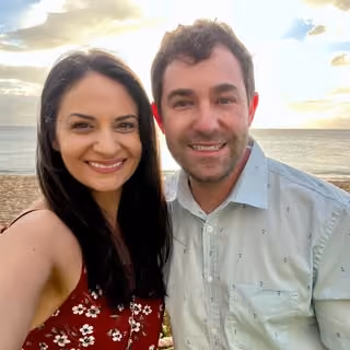 Smiling man and woman posing for a selfie by the ocean with a cloudy sky at sunset.