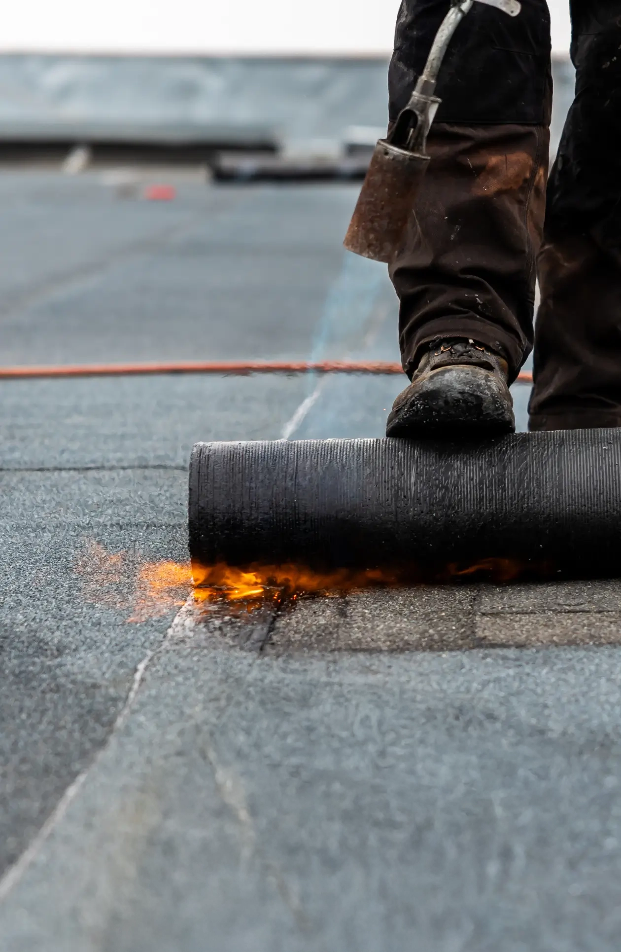 Worker applying heated bitumen roofing membrane with torch, sealing waterproof layer for durable flat‑roof protection.
