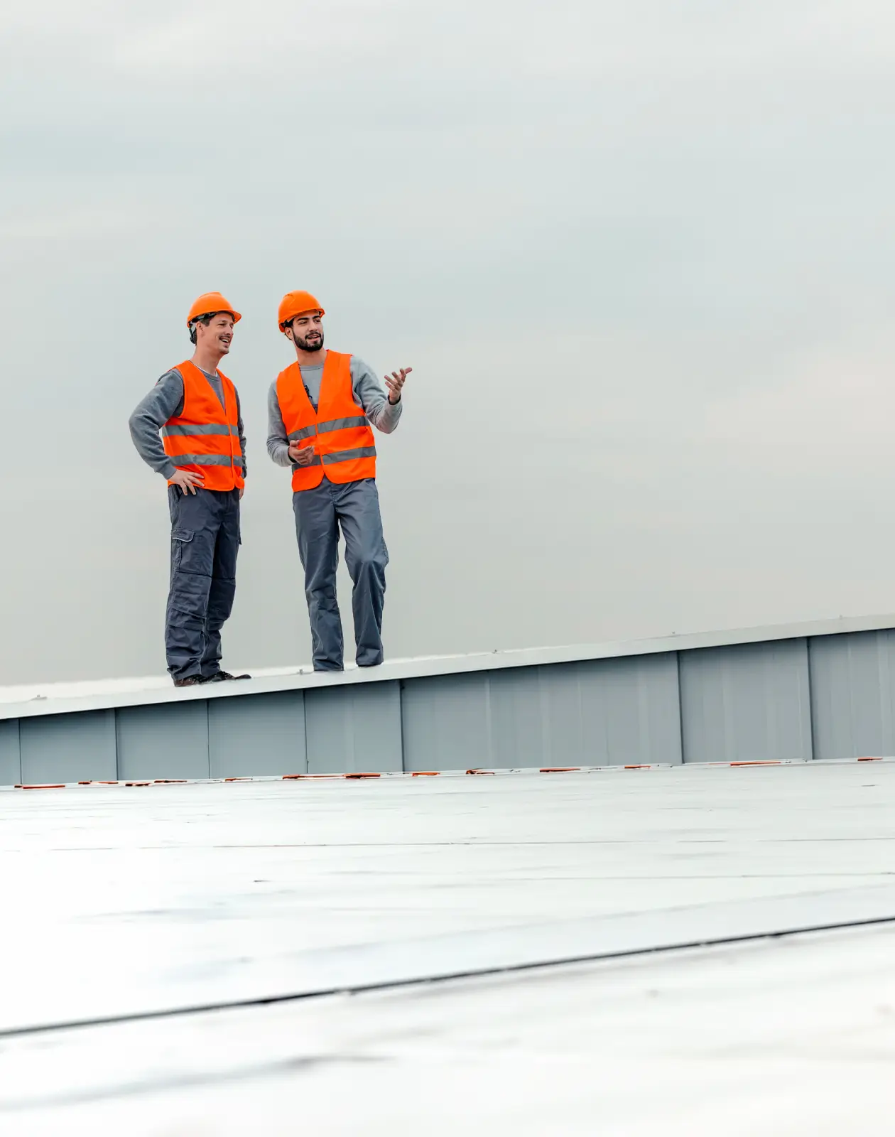 Two construction professionals in orange safety vests and helmets stand on a metal roof, discussing inspection results under an overcast sky.