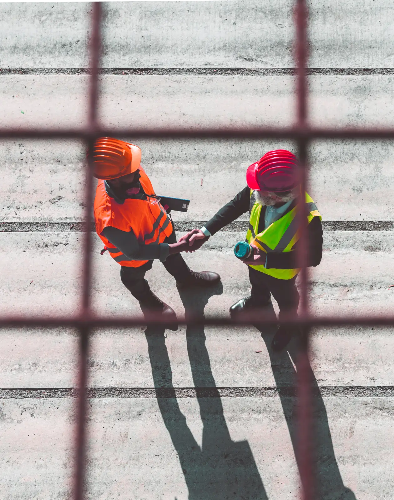 Two construction professionals wearing safety vests and helmets shake hands on a concrete surface, symbolizing agreement and project collaboration.
