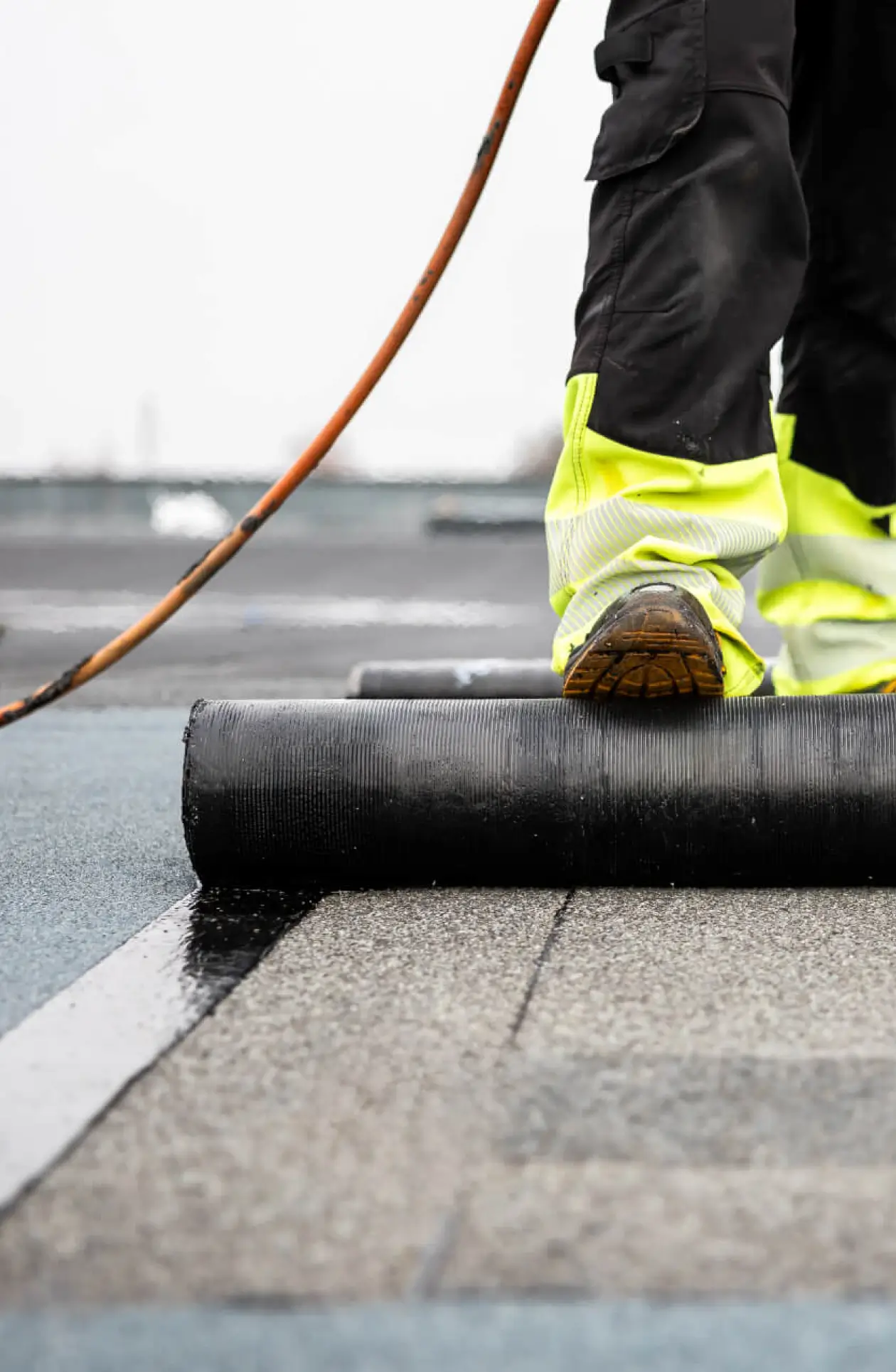 Worker in high‑visibility pants applying heated bitumen membrane on a flat roof, illustrating durable waterproofing for commercial roofing systems.
