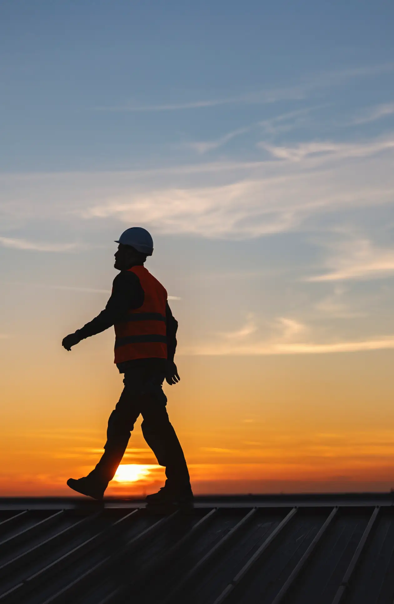 Silhouette of a construction worker in safety gear walking across a rooftop at sunset, symbolizing proactive maintenance and long‑term roof protection.