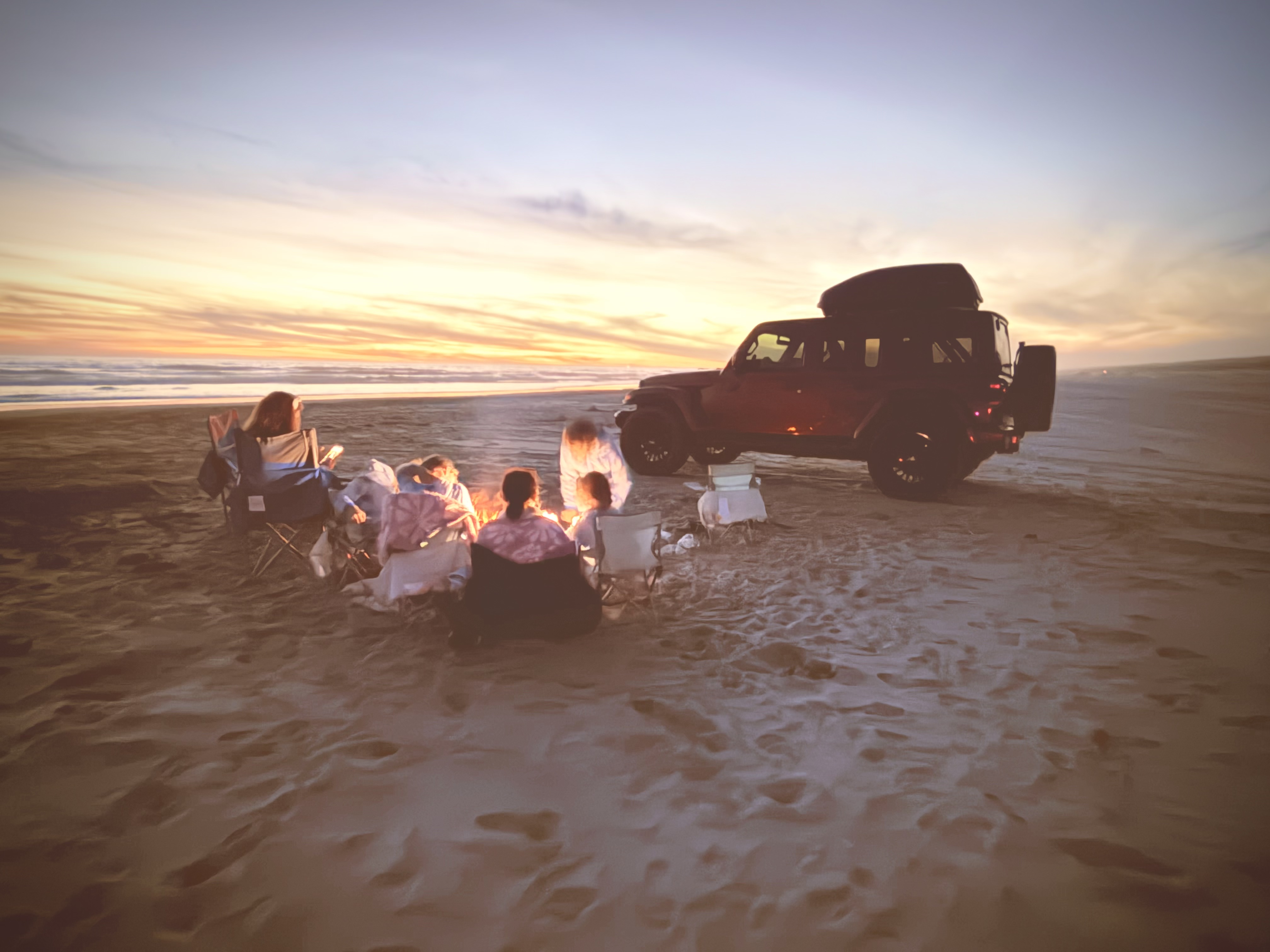 Family around a beach campfire at sunset next to a red Jeep