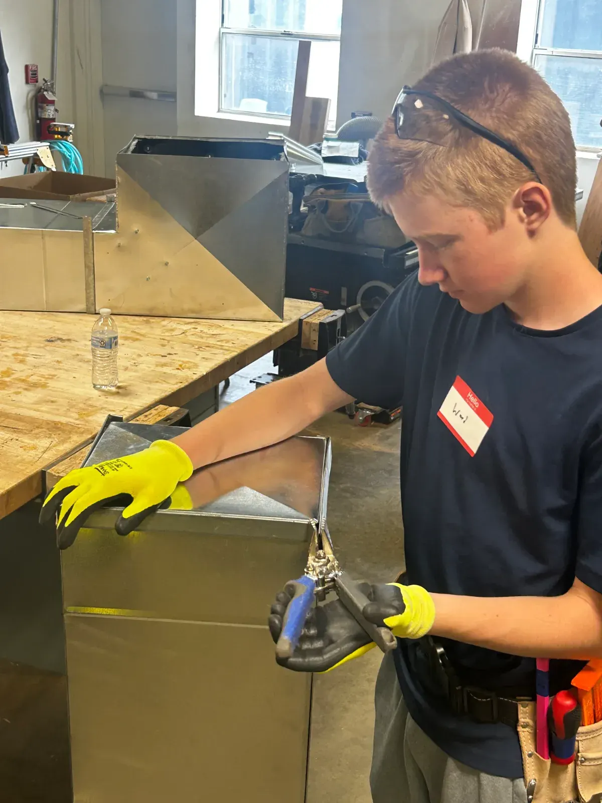 A student in safety glasses and gloves shaping sheet metal at a bench