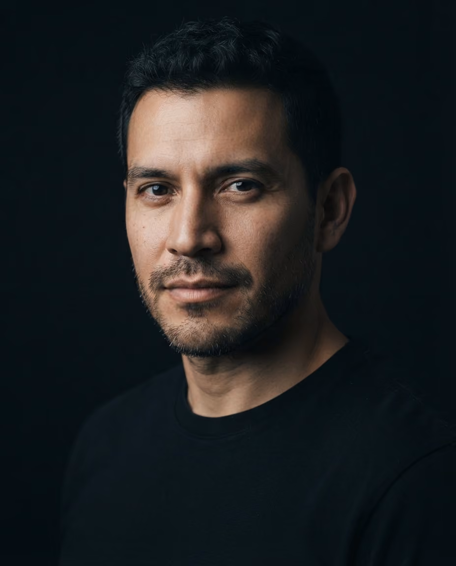 Close-up portrait of a man with short dark hair and light stubble wearing a black shirt against a dark background.