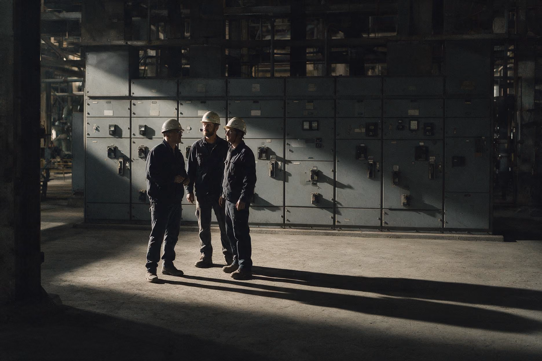 Three workers in helmets standing and talking inside an industrial facility with control panels.