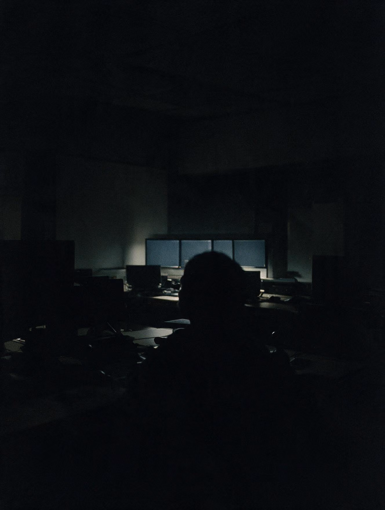 Silhouette of person in dark room with multiple computer monitors lit on a desk.