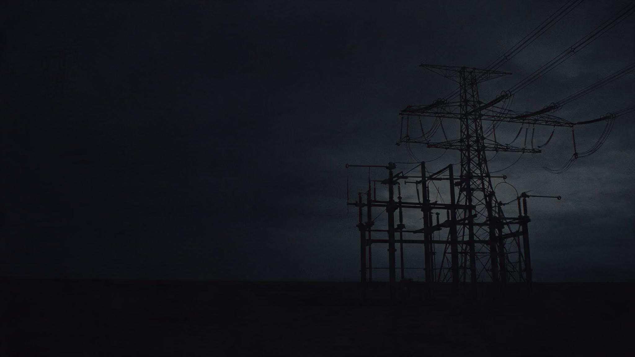 Silhouette of an electrical transmission tower and power lines against a dark, cloudy sky.