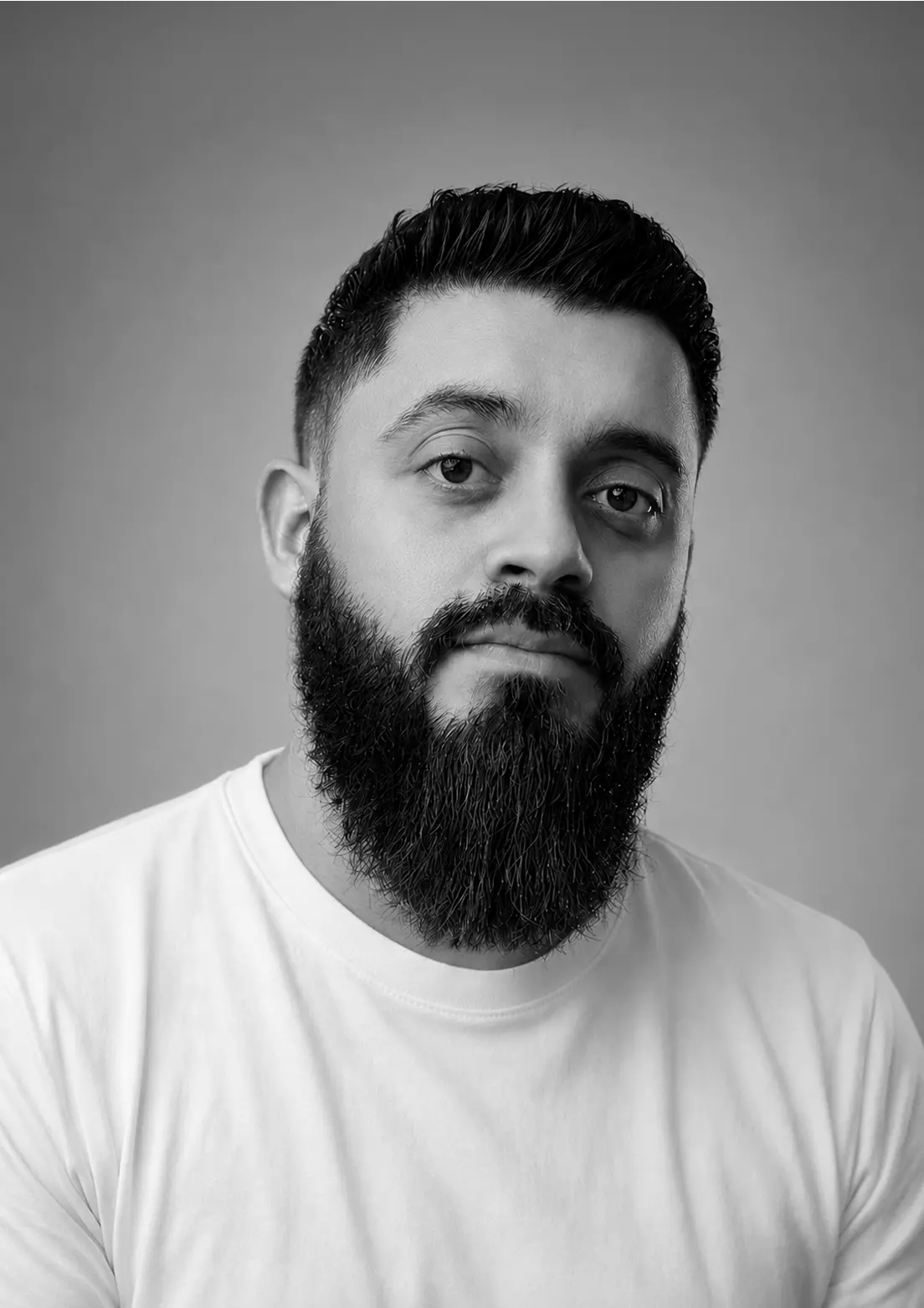 Portrait en noir et blanc d'un homme avec une barbe épaisse et les cheveux coiffés en arrière, portant un t-shirt blanc, regardant directement vers l'appareil photo sur un fond uni gris.