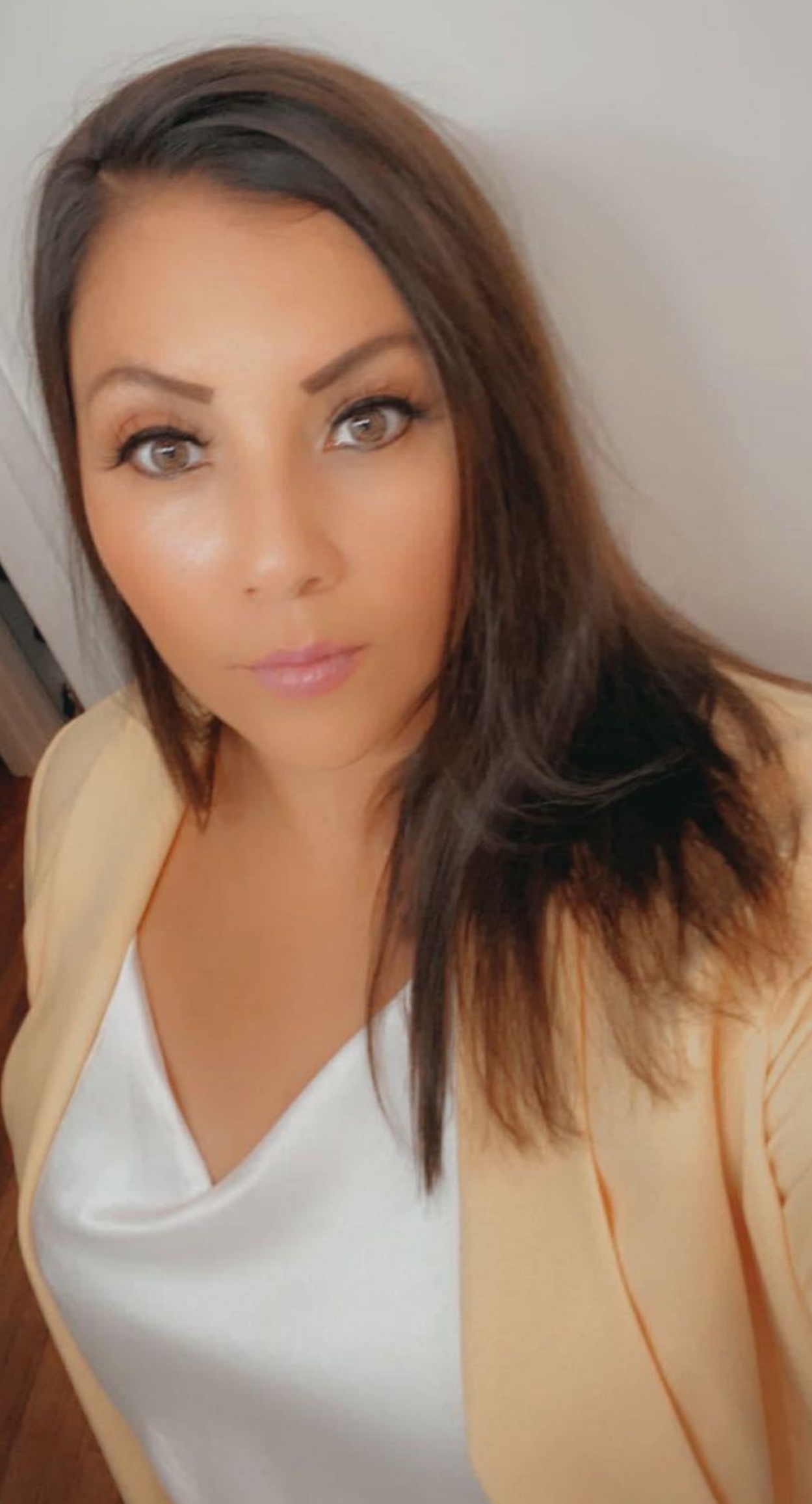 Close-up of a woman with long dark hair wearing a light yellow blazer over a white top, looking directly at the camera with a neutral expression.
