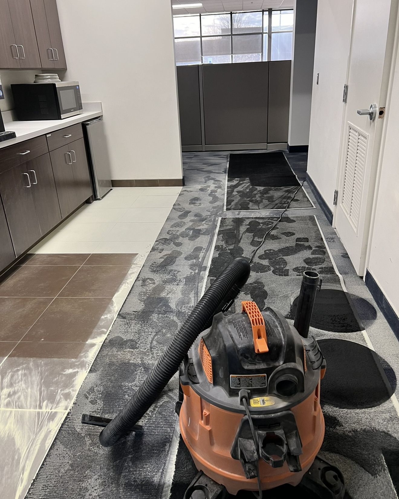 An orange and black wet-dry vacuum cleaner sitting on a carpeted office floor with visible wet footprints near office cubicle partitions, next to a kitchen area with dark cabinets, microwave, mini fridge, and tiled flooring partially covered with plastic sheets.