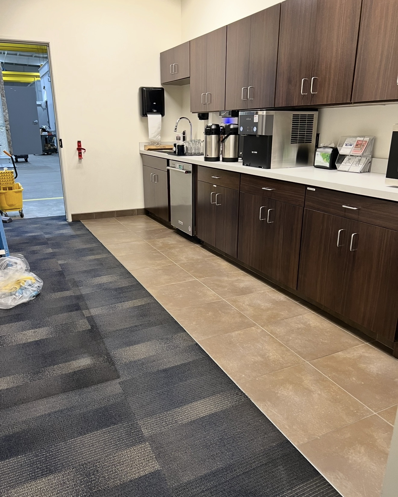 Modern break room kitchen area with dark wood cabinets, white countertop, stainless steel dishwasher, coffee dispensers, and an ice machine, adjacent to a door leading to an industrial space with yellow mop bucket visible.