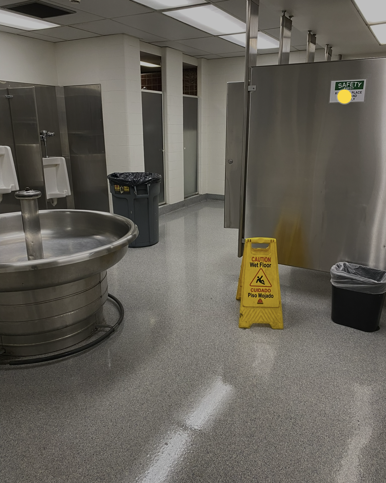 Interior of a public restroom with stainless steel fixtures, including a round communal sink, urinals on the left, and toilet stalls with metal doors on the right; a yellow caution sign warns of a wet floor near a trash container, and another trash bin is against the back wall.