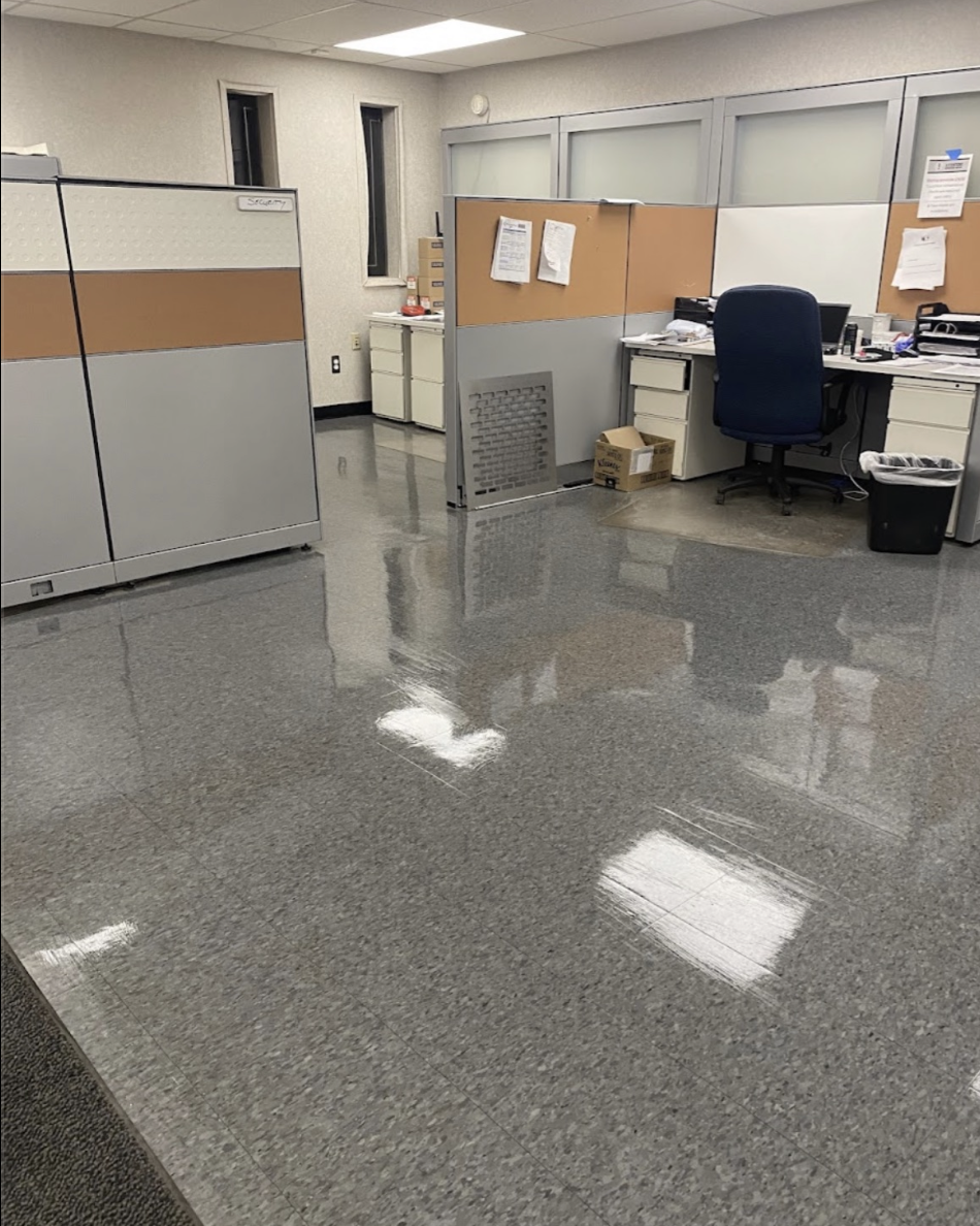 Office cubicle area with gray and brown partition walls, a desk with papers and office supplies, a blue office chair, a cardboard box, and a black trash bin on shiny gray tiled floor reflecting overhead lights.