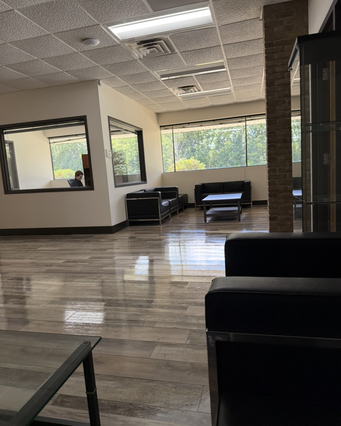 Modern office waiting area with shiny wood-look flooring, black leather chairs and sofas around low tables, large windows showing trees outside, and a man sitting and working alone in a glass-walled office room.