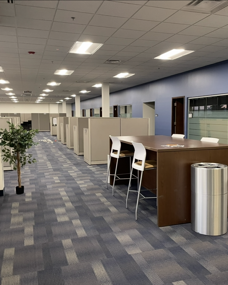 Modern office space with rows of beige cubicles along a blue patterned carpet, a wooden high table with white chairs on the right, a silver trash can beside the table, and a potted plant on the left under bright ceiling lights.