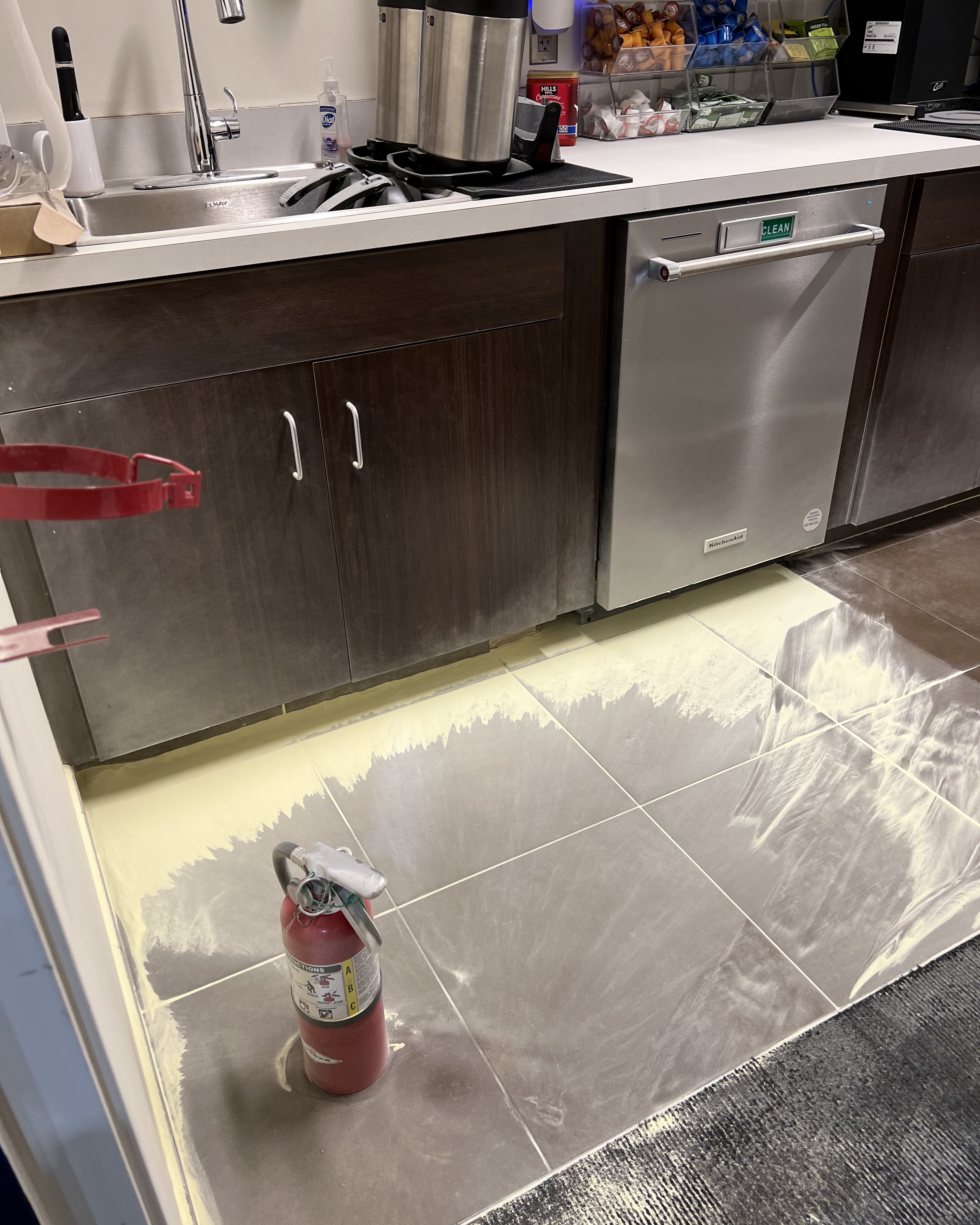 Kitchen area with dark wood cabinets, stainless steel dishwasher labeled clean, sink with soap dispenser, and a coffee station on the counter; the tiled floor is covered with a powdery yellow substance spread around a red fire extinguisher.