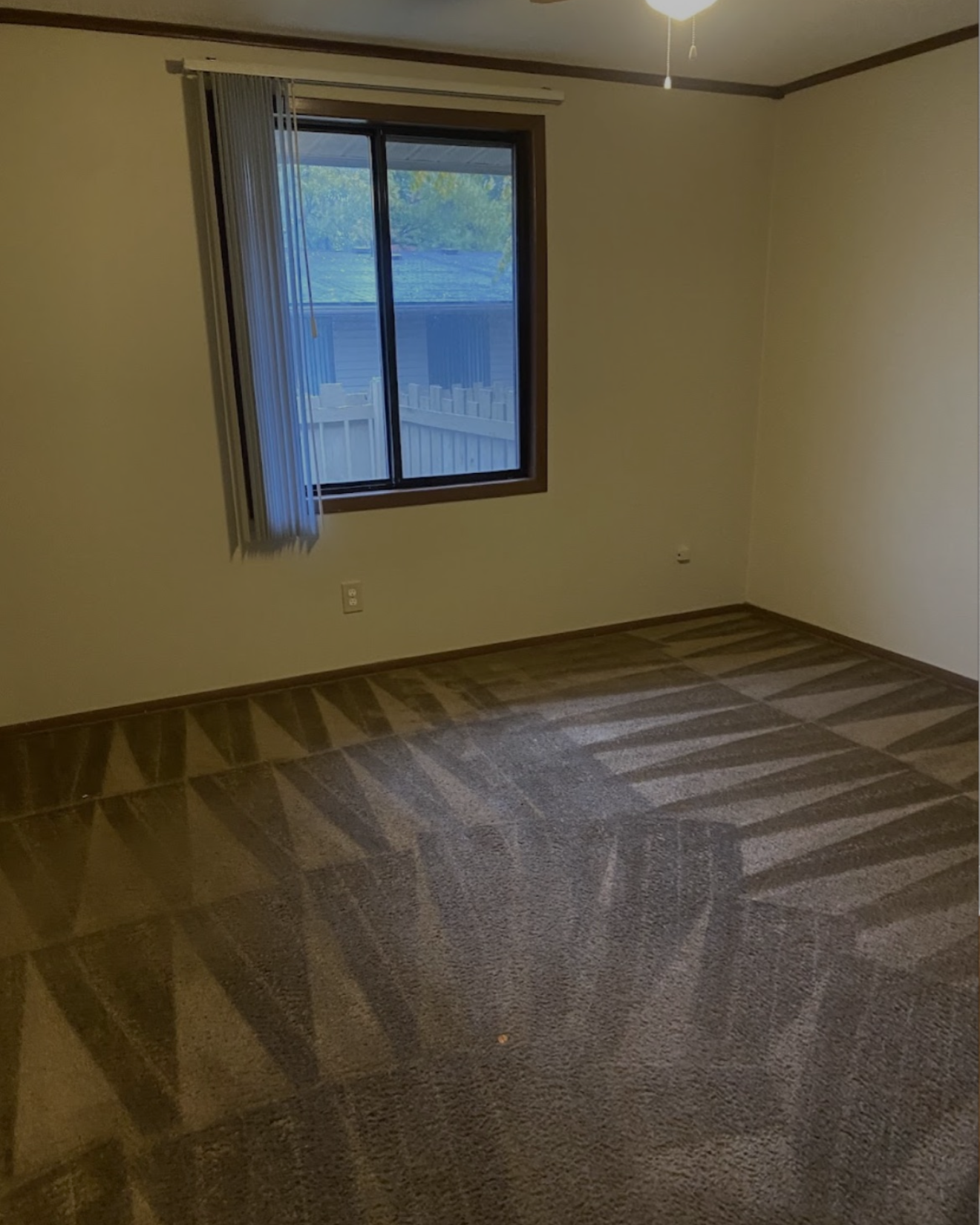 Empty room with light beige walls, brown carpet with vacuum lines, a single window with partially open vertical blinds showing an outdoor fence and roof, and ceiling fan light on.