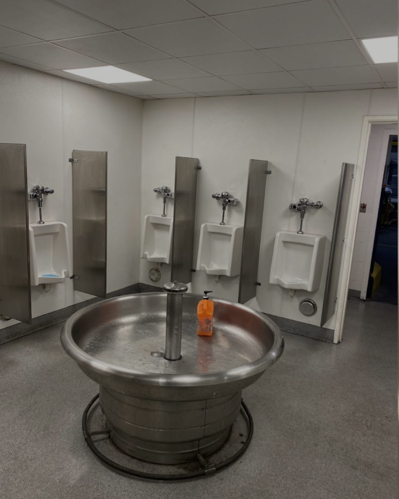 Public restroom with four white urinals mounted on a white wall, each separated by metal privacy dividers, and a large round stainless steel basin sink with an orange soap dispenser in the center of the room.