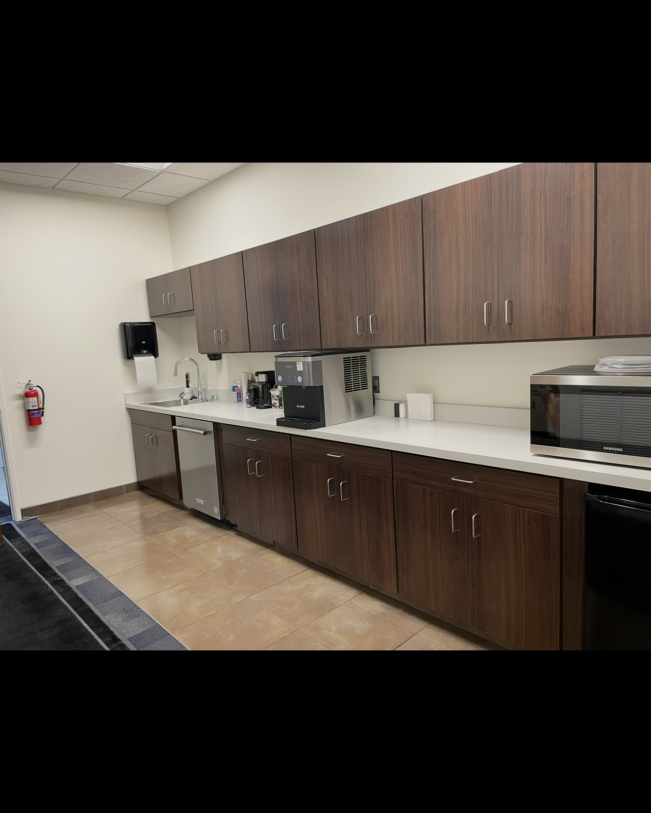 A modern office kitchenette with dark wood cabinets, white countertops, stainless steel dishwasher, coffee maker, a microwave, and a sink with a paper towel dispenser on the wall, with a fire extinguisher mounted near the doorway.