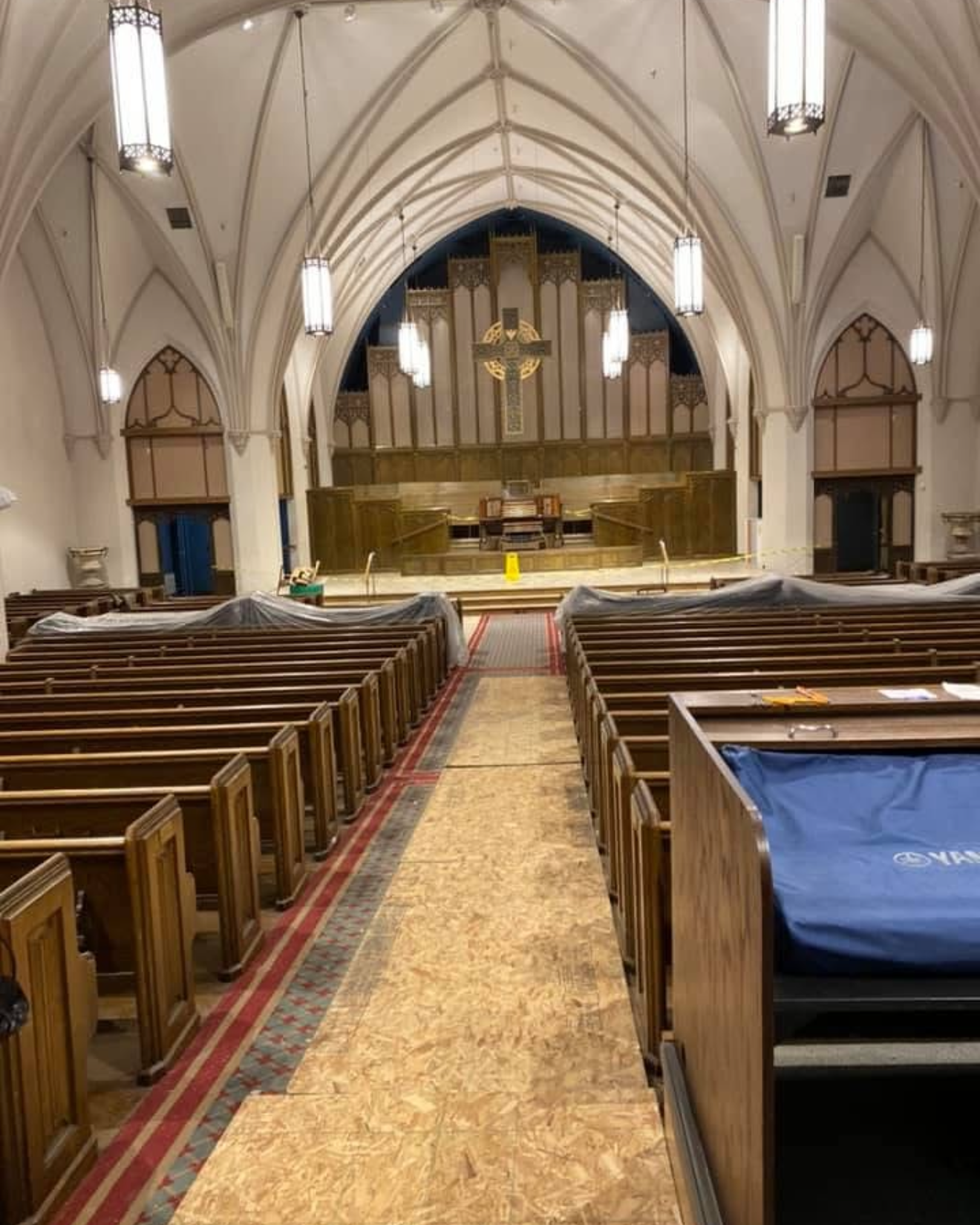 Interior of a church with wooden pews on both sides of a central aisle covered with plywood boards, leading to a raised altar with a large decorative cross on the back wall, gothic-style arches on the ceiling, and hanging lights. Some pews near the front are covered with plastic sheets.