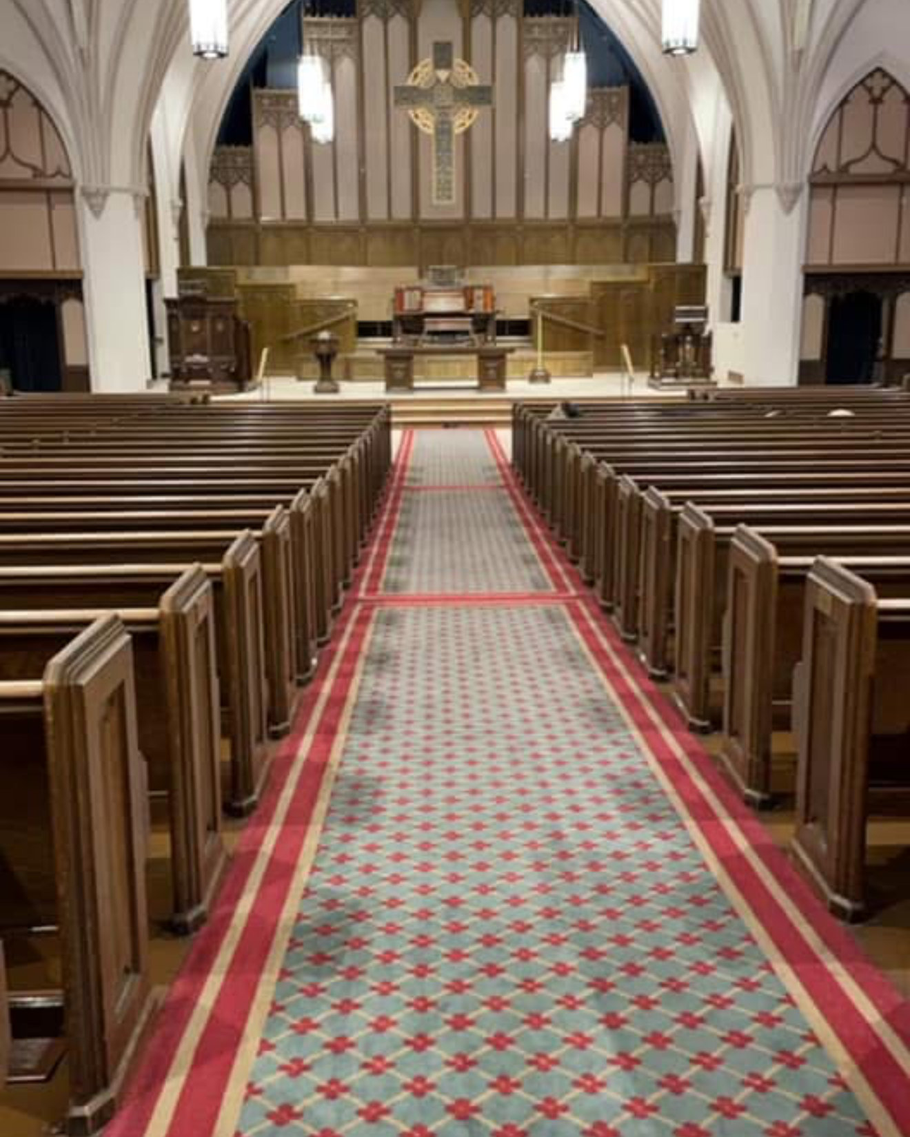Interior view of a church with wooden pews on both sides of a patterned carpeted aisle leading to an altar, ornate wooden paneling, and a large decorative cross on the wall behind the altar under vaulted ceilings with hanging lights.