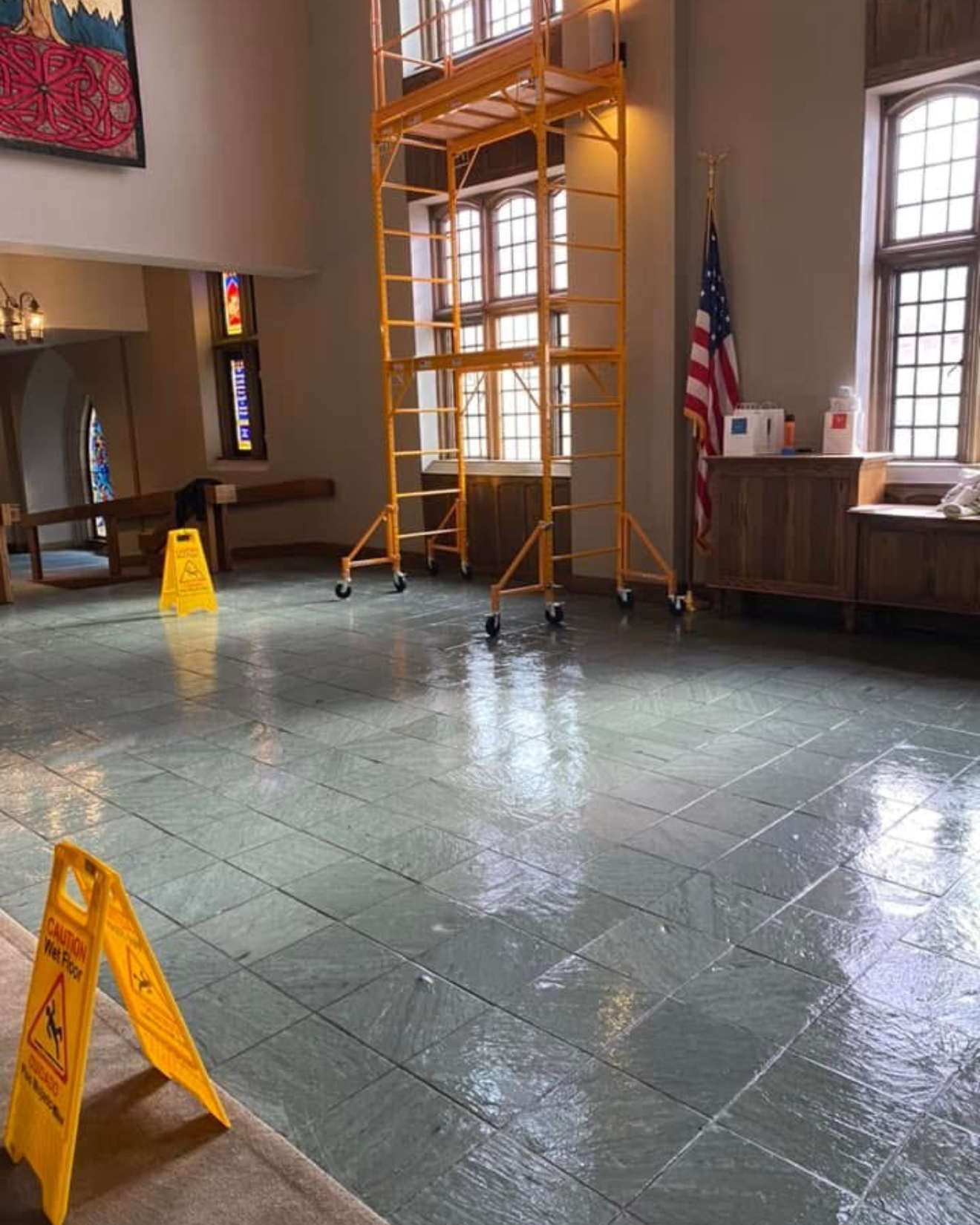 Indoor room with wet shiny floor marked by two yellow wet floor caution signs, a large yellow scaffold on wheels near windows, and an American flag next to a wooden cabinet.