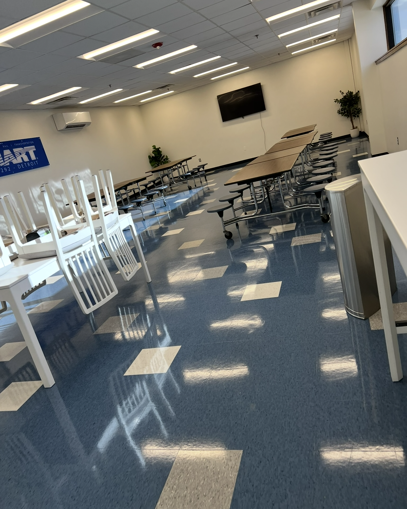 Empty cafeteria or break room with blue and white tiled floor, long tables with attached black stools on wheels, white chairs stacked upside down on white tables, a wall-mounted TV, two potted plants, and a blue sign reading 'SMART 292 - Detroit'.
