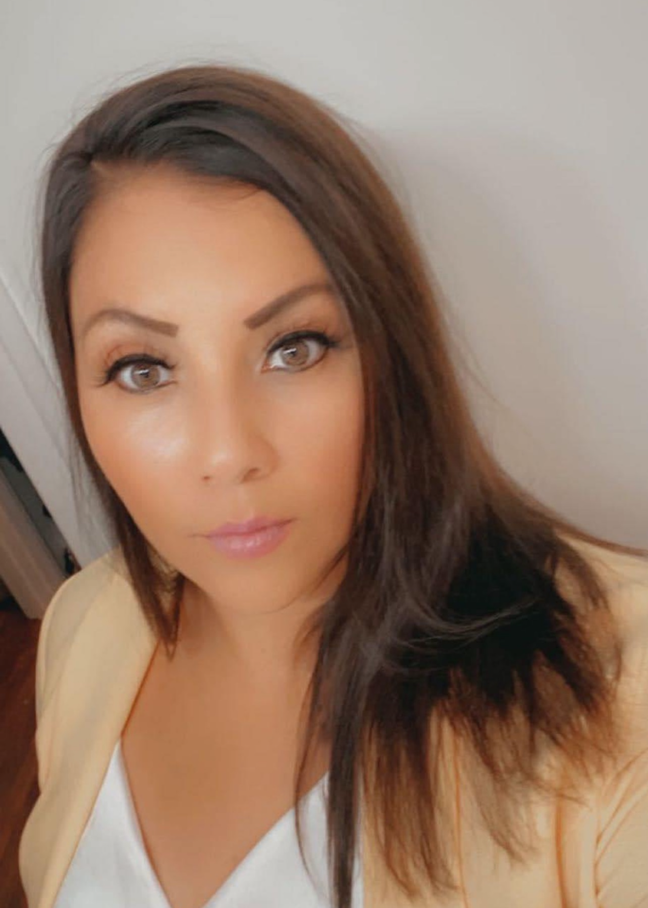 Close-up of a woman with long dark brown hair, wearing a light yellow jacket over a white top, looking directly at the camera with a neutral expression against a plain light-colored background.