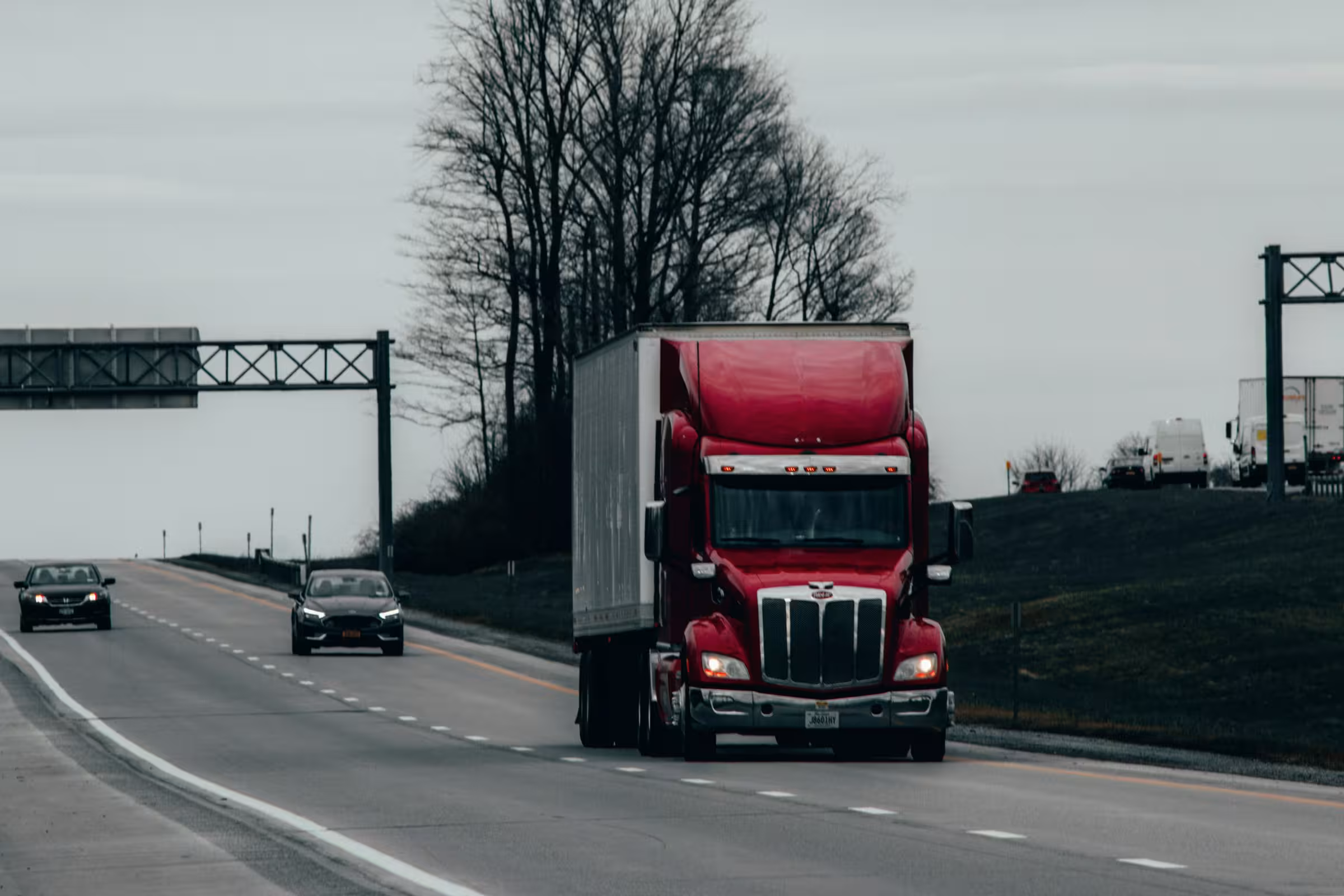 Red semi truck driving on a multi-lane highway with cars in adjacent lanes and leafless trees along the roadside under an overcast sky.