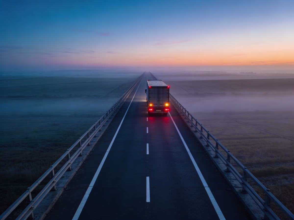 A truck with red taillights illuminated drives on a long, empty highway at dawn, surrounded by foggy fields under a blue and orange sky.