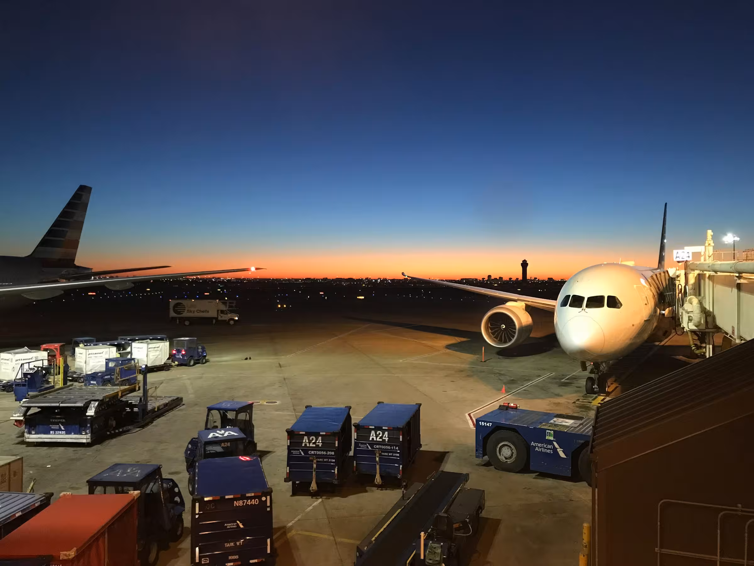 American Airlines plane parked at the gate on the tarmac at dawn with airport ground service vehicles and cargo containers nearby, under a clear sky transitioning from orange at the horizon to deep blue above.
