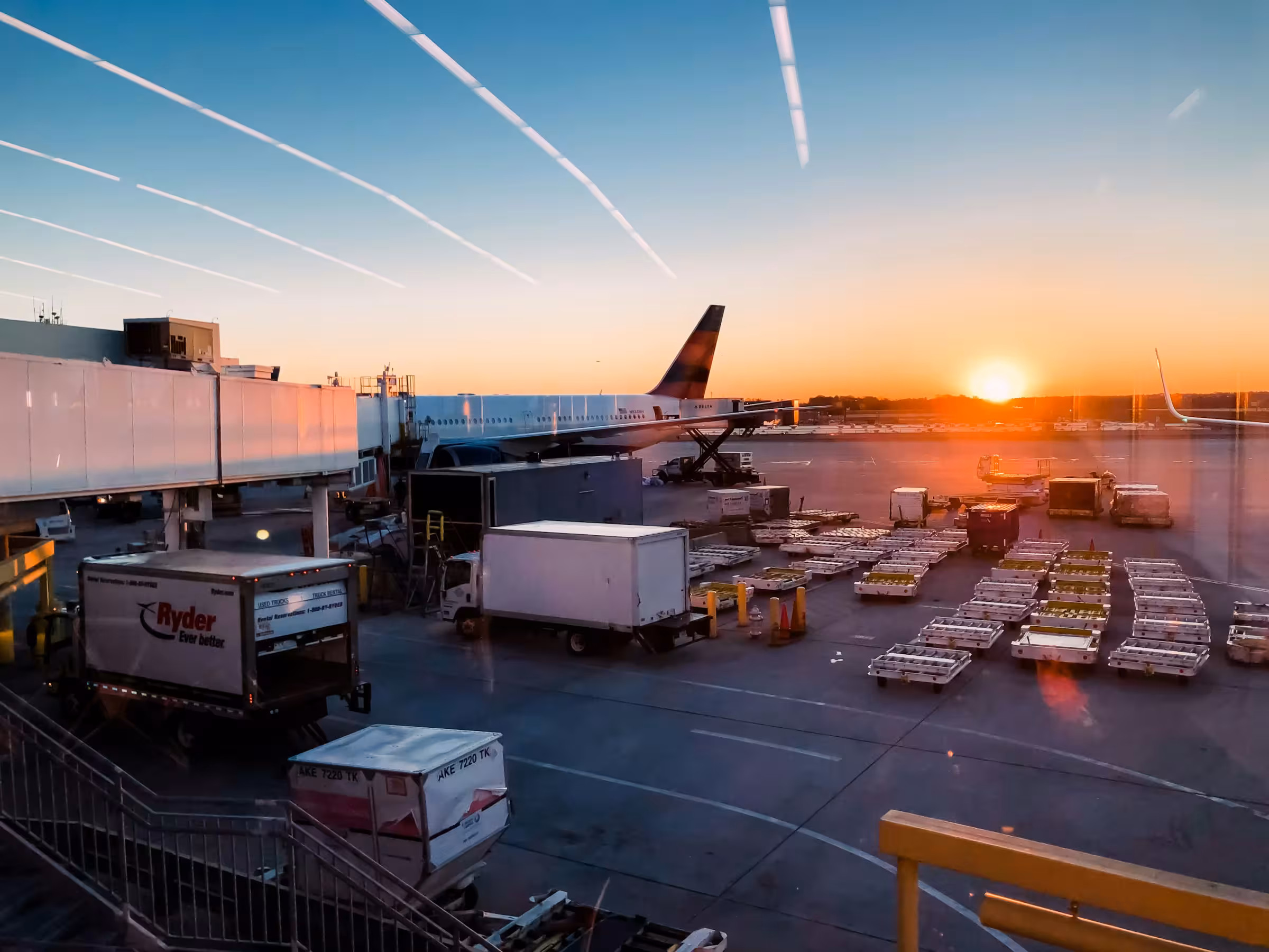Airport tarmac at sunset with a large commercial airplane parked at a gate, surrounded by cargo transport carts and vehicles under a partly cloudy sky.