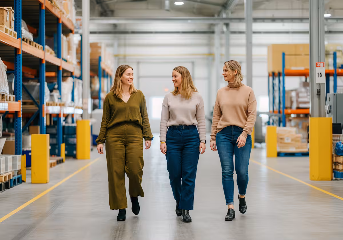 Three women walking and smiling inside a spacious warehouse with shelves stocked with boxes on both sides.