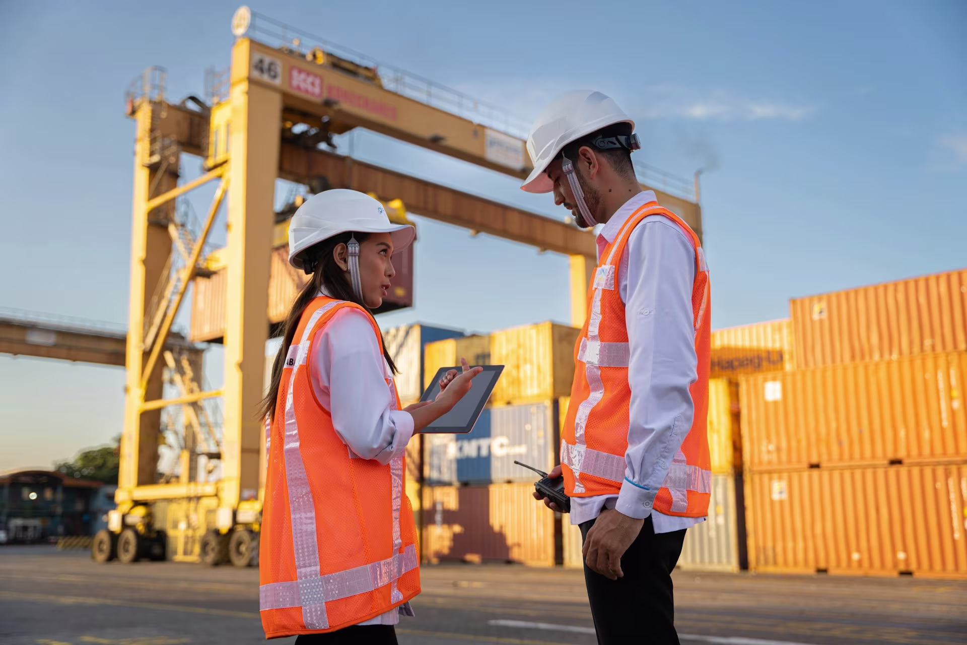 Two logistics workers in safety vests and hard hats discussing work using a tablet at a shipping container terminal