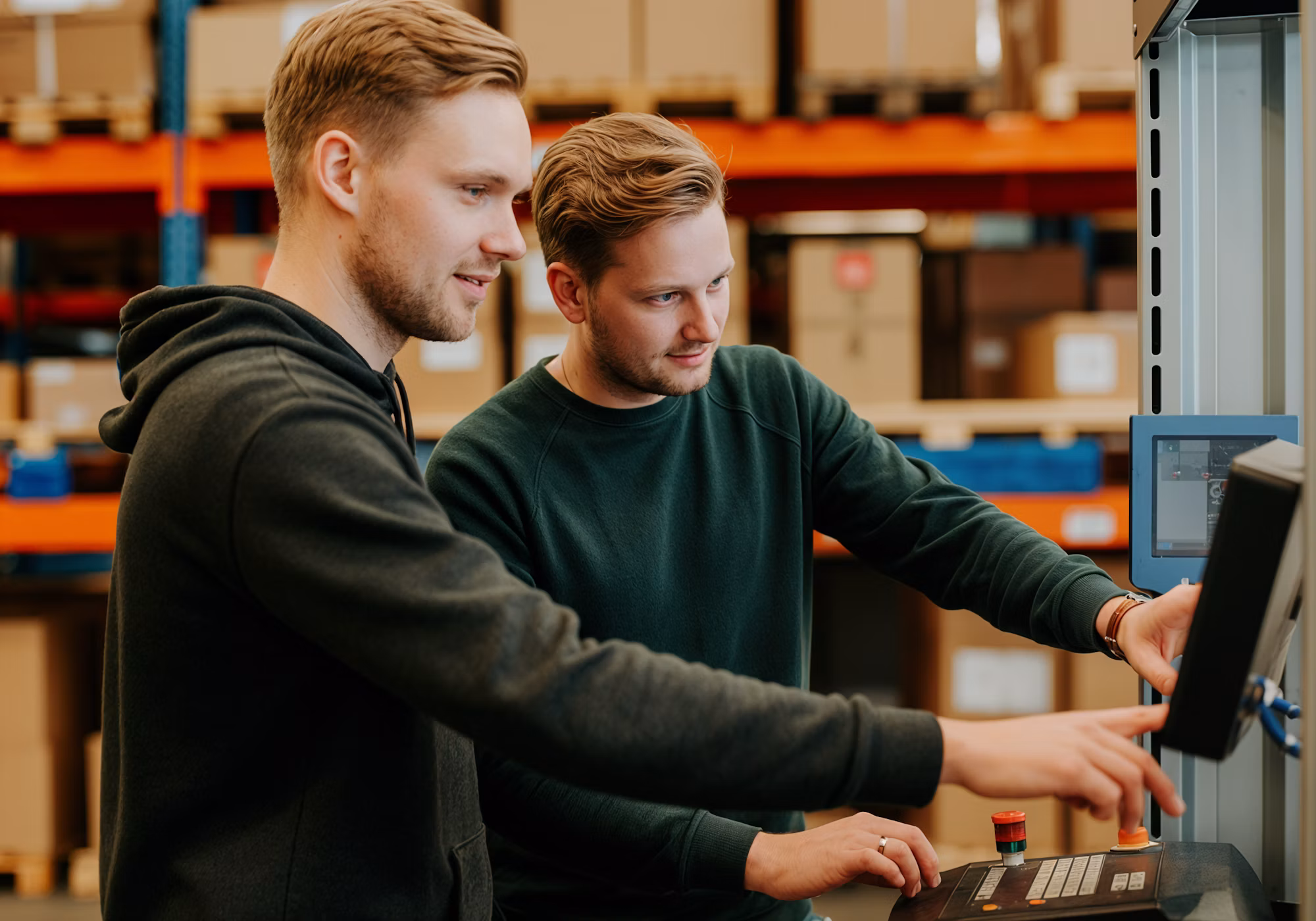 Two male warehouse operators working together to configure settings on a touch screen control panel of industrial machinery