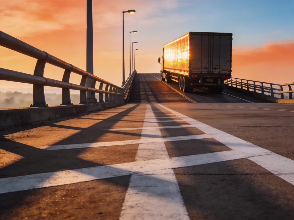 Rear view of a cargo truck driving up an elevated bridge under a warm sunset sky