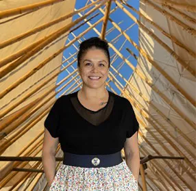 Smiling woman with dark hair standing in a wooden frame structure under blue sky.