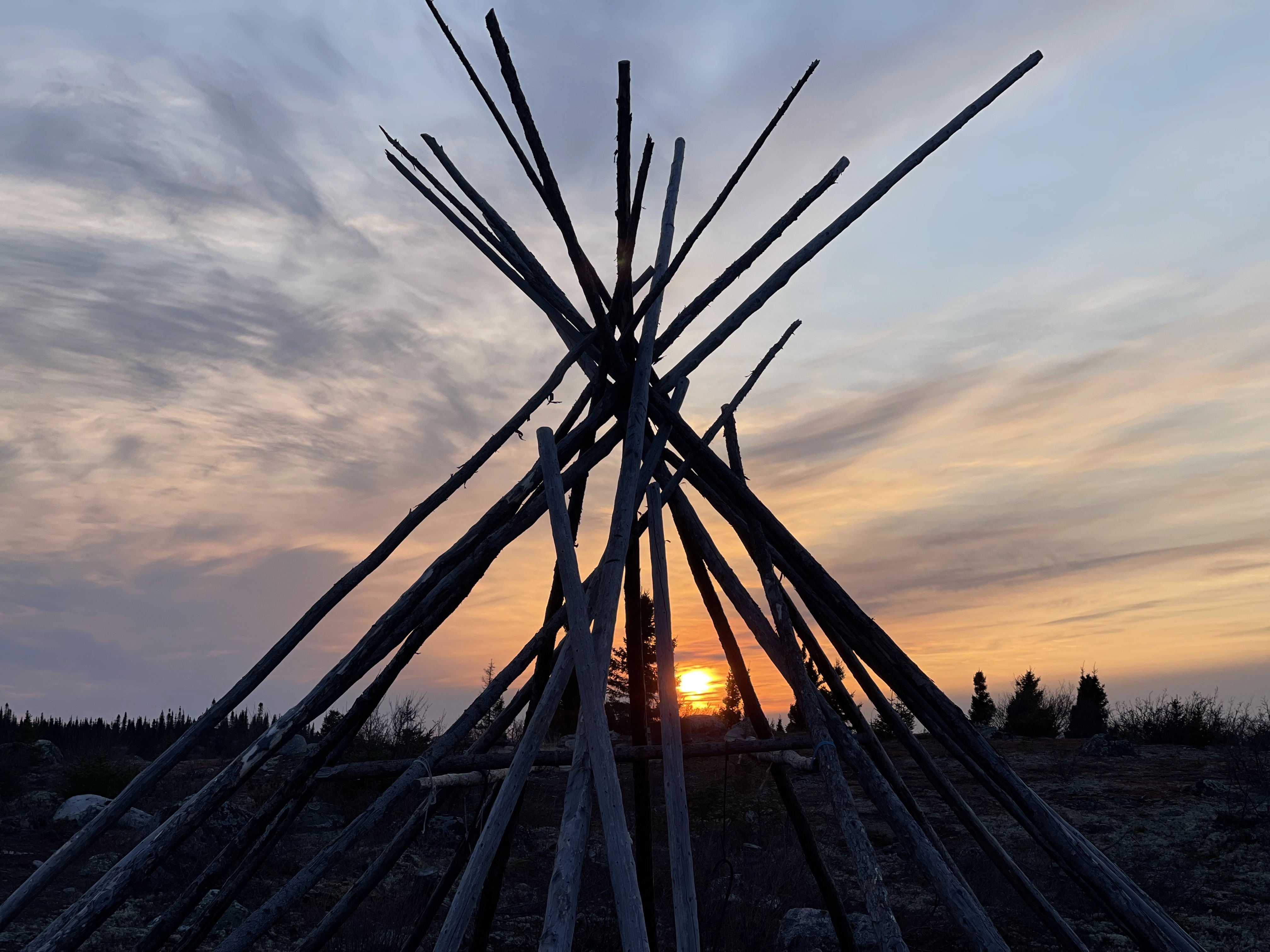Wooden teepee frame silhouetted against a colorful sunset sky with wispy clouds.